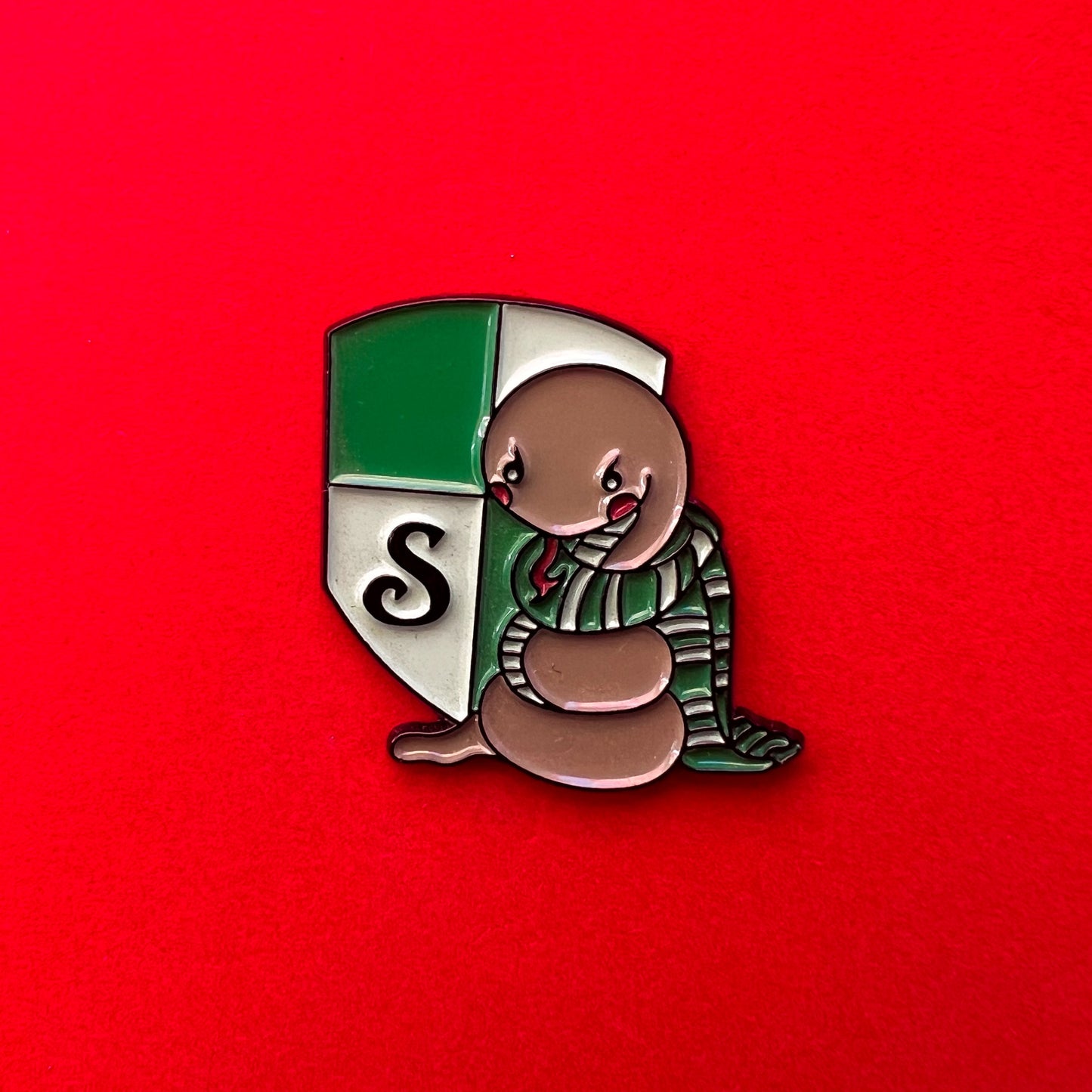 Pin de Slytherin con una serpiente tierna sobre el escudo de la casa, en tonos verdes y plateados.